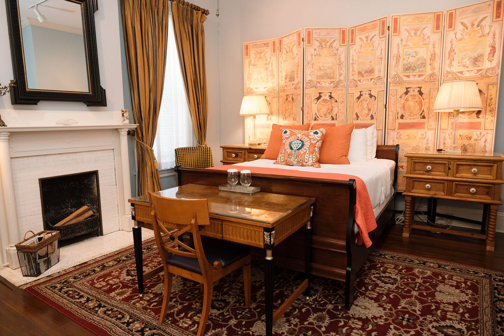 Bedroom suite featuring a wooden sleigh bed with orange accents, a decorative folding screen, antique desk, and white fireplace.