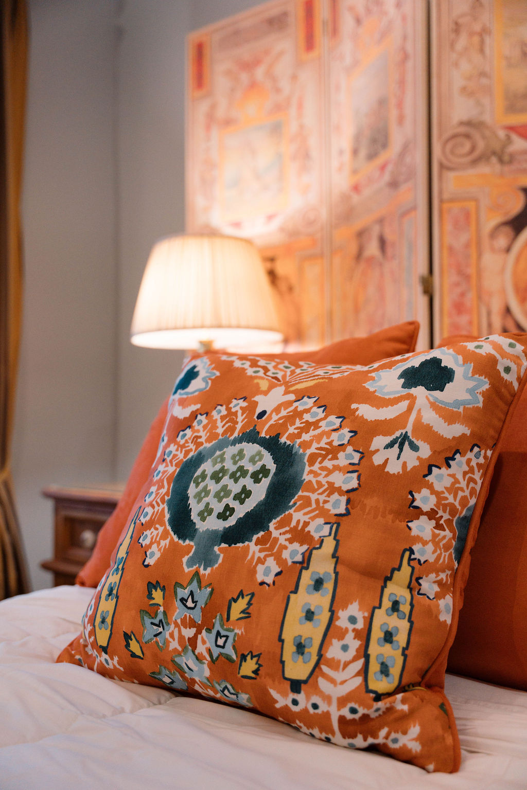 Close-up detail of an orange embroidered throw pillow with teal and white floral patterns on a bed, set against a soft-focus background of a lamp and decorative screen.