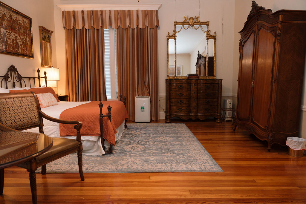 Spacious bedroom suite featuring hardwood floors, a large wooden armoire, an antique dresser with mirror, and a bed with an orange throw blanket.