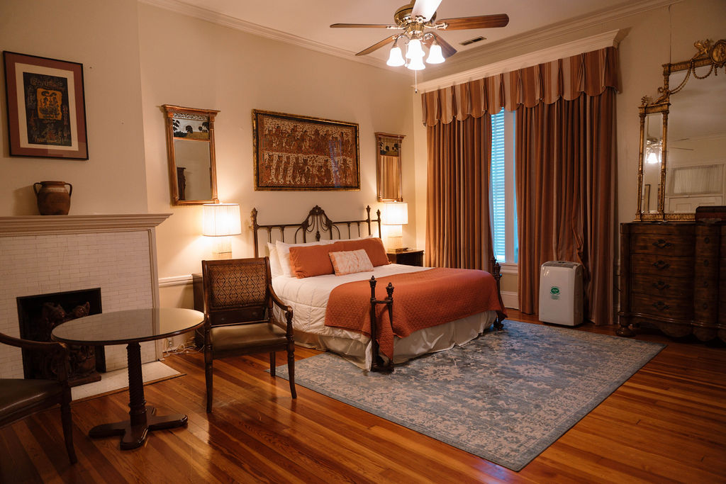 Spacious bedroom suite featuring hardwood floors, an iron bed with orange accents, a sitting area with a fireplace, and a large wooden dresser.