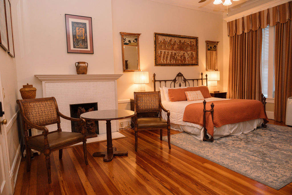 Bedroom suite featuring gleaming hardwood floors, a sitting area with two chairs by a white fireplace, and an iron bed with orange accents.