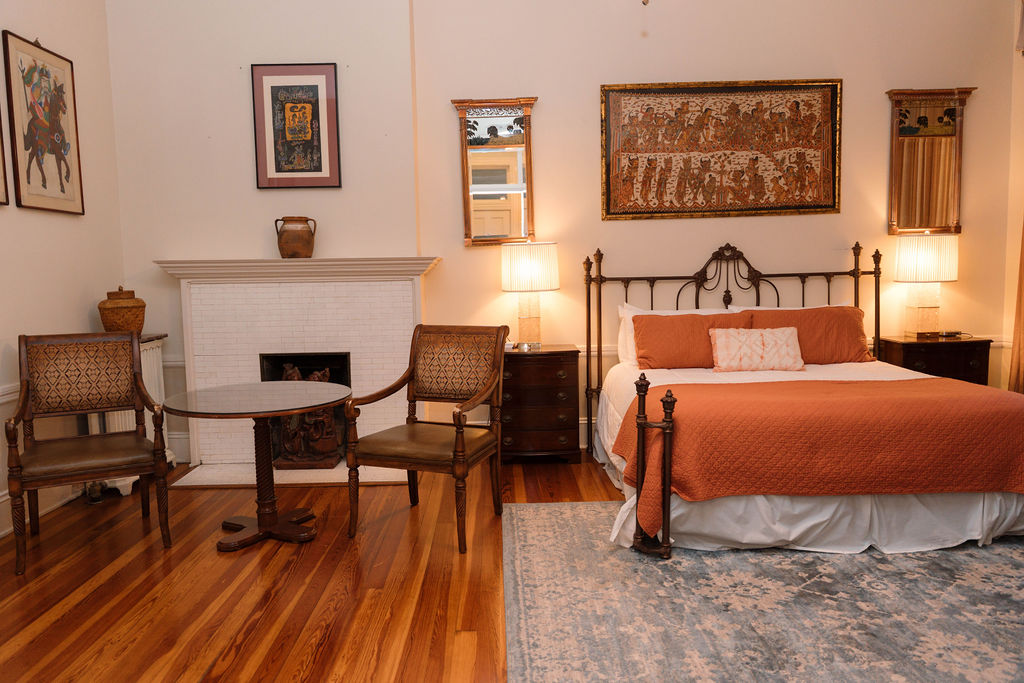 Bedroom suite featuring polished hardwood floors, a sitting area with two chairs by a white fireplace, and a black iron bed with orange bedding.