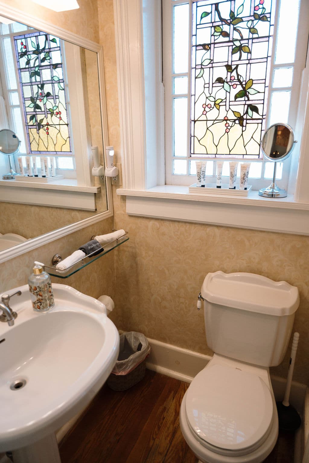 Ensuite bathroom featuring a white pedestal sink, large mirror, and a window with intricate floral stained glass above the toilet.