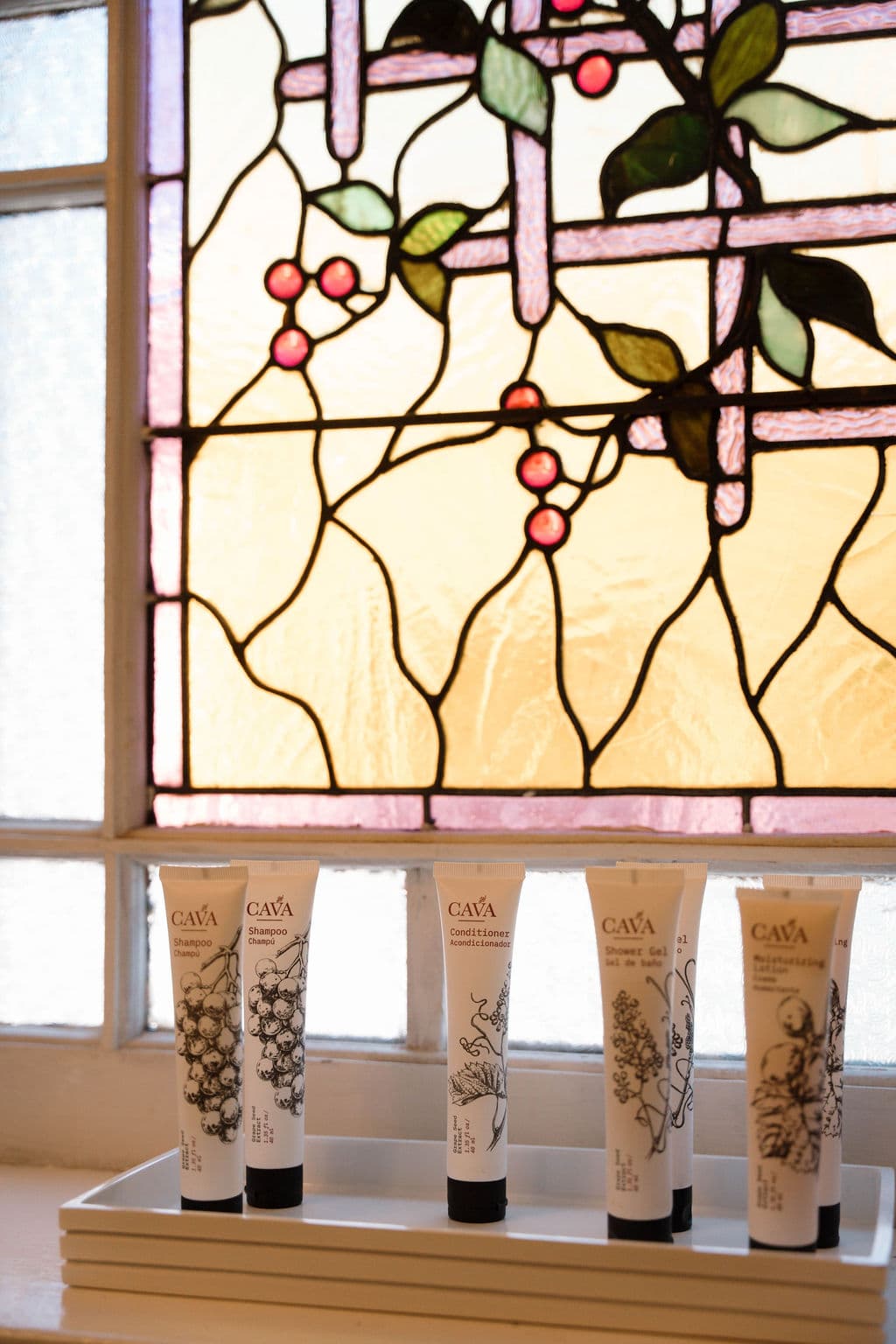 Close-up detail of premium bath amenities arranged on a windowsill in front of a stained glass window featuring a floral vine and berry motif.