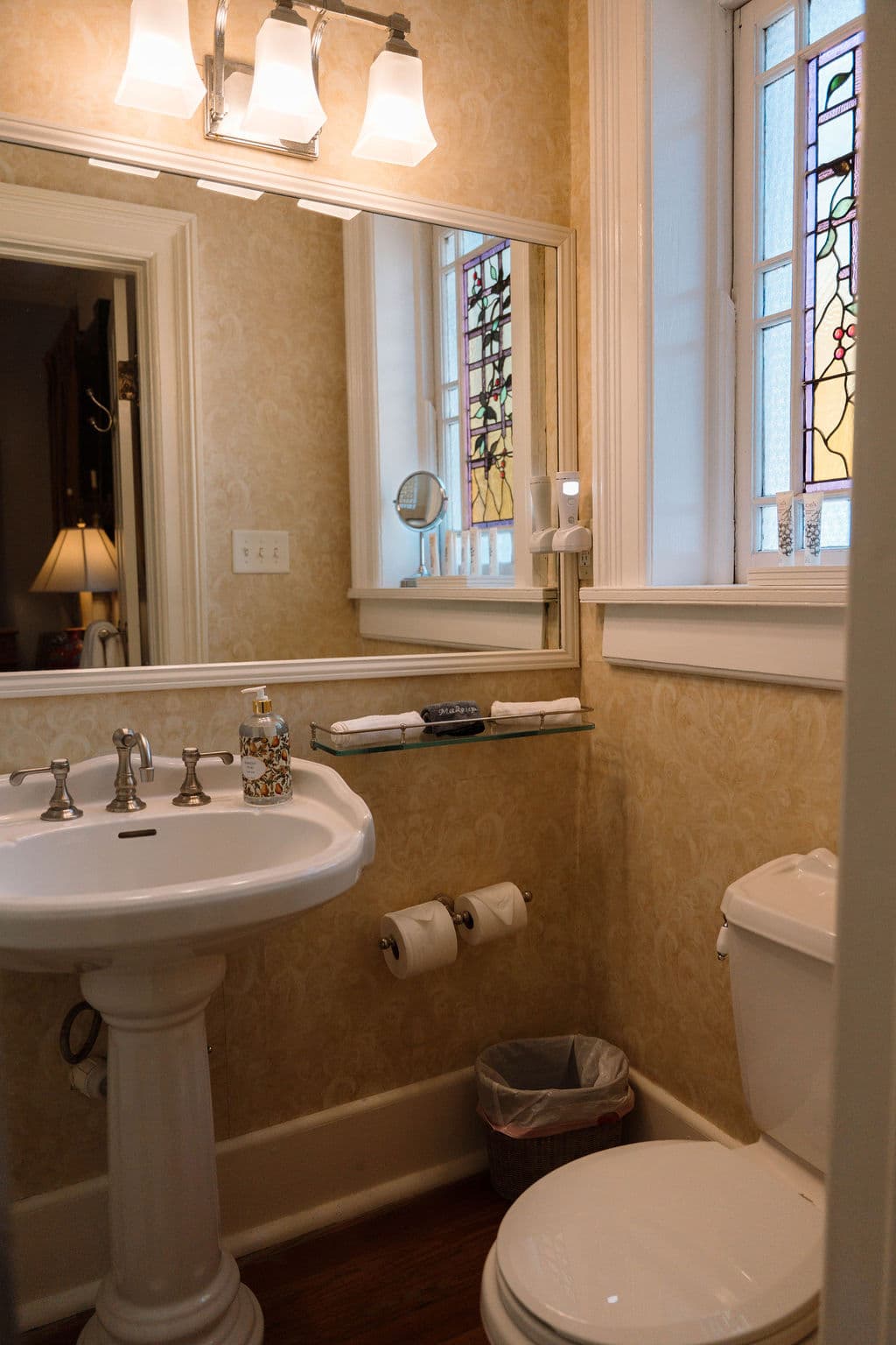 Bathroom suite featuring a white pedestal sink, vanity mirror with light fixture, and a decorative stained glass window reflecting in the glass.