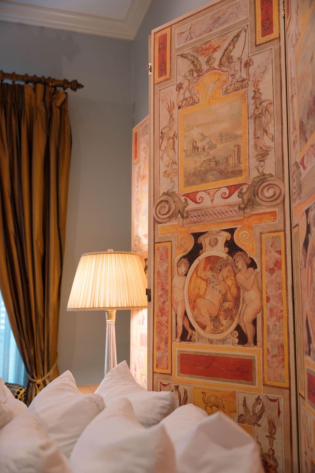 Close-up detail of a bedroom featuring plush white pillows, a lit bedside lamp, and an intricate, hand-painted decorative screen with gold drapes in the background.