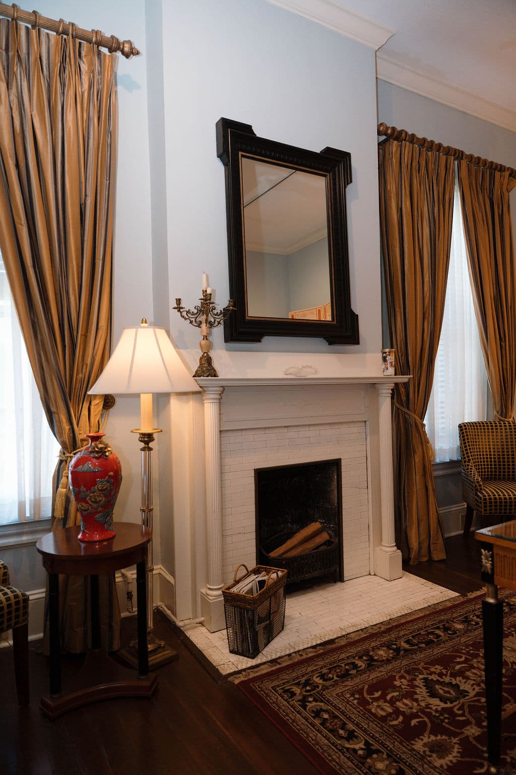 White fireplace mantel with a dark framed mirror, flanked by tall windows with gold drapes and a floor lamp.