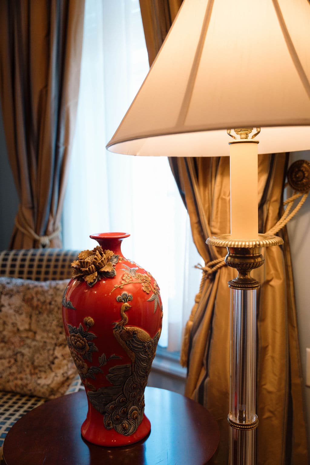 Close-up of a red decorative vase with gold relief details sitting on a side table, illuminated by a floor lamp against a backdrop of gold drapes and a patterned armchair.