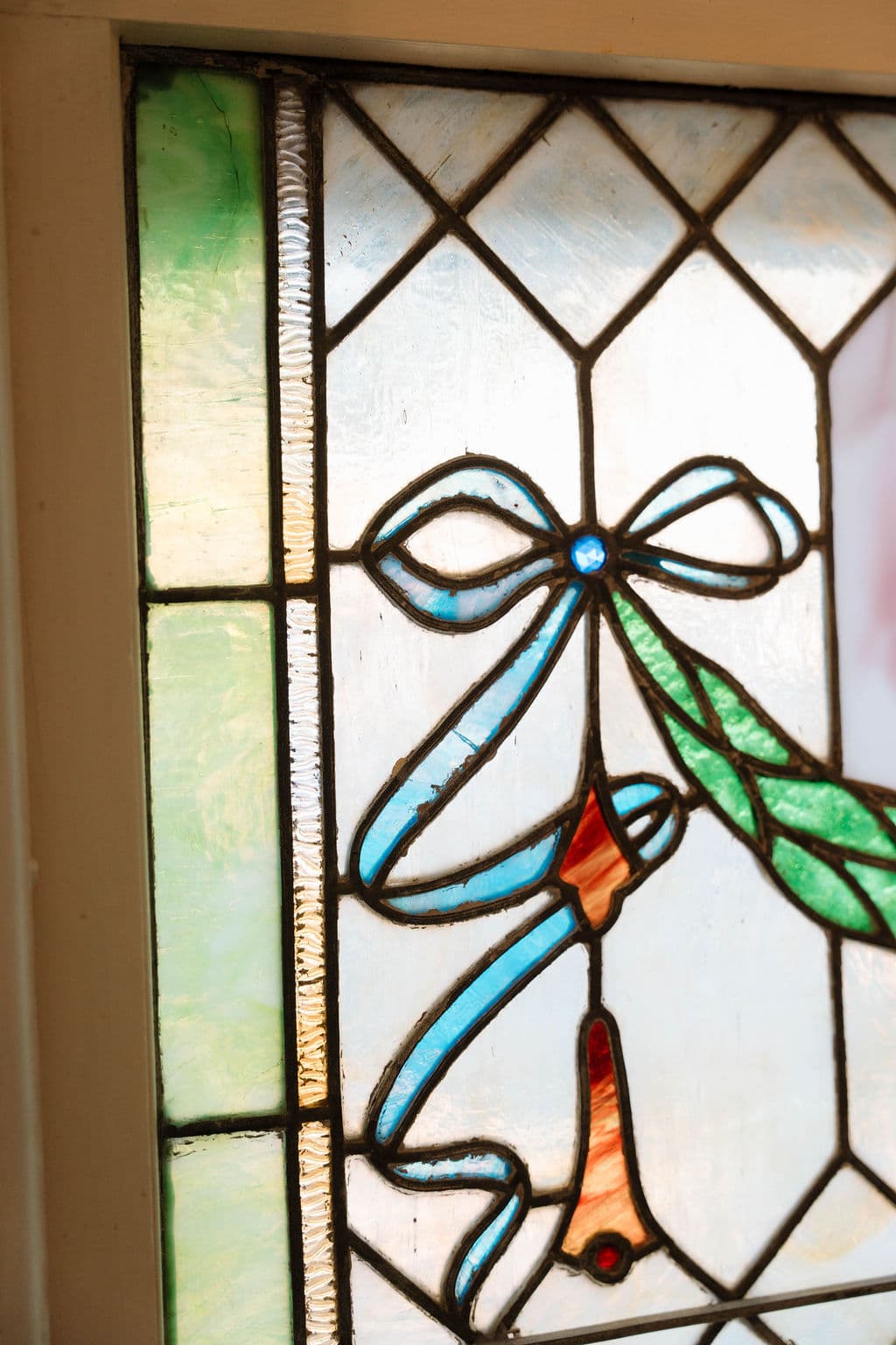 Close-up detail of a stained glass window featuring a blue ribbon bow, green foliage, and a hanging tassel motif set within a geometric diamond pattern.