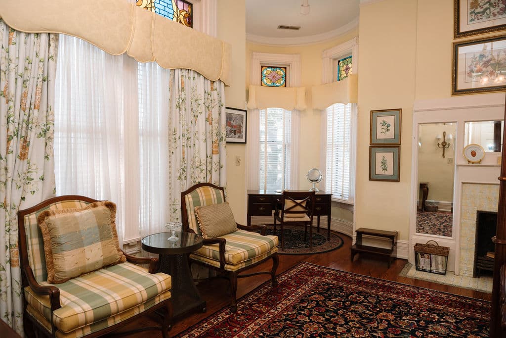 Sunny sitting area featuring two striped armchairs, large windows with floral drapes, a Persian rug, and a writing desk nestled in a bay window.