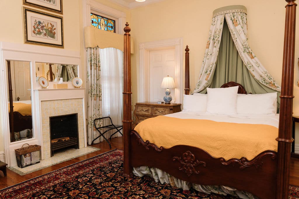 Bedroom suite featuring a dark wood four-poster bed with a floral canopy, a yellow throw blanket, a white fireplace with a mirror, and a Persian rug.