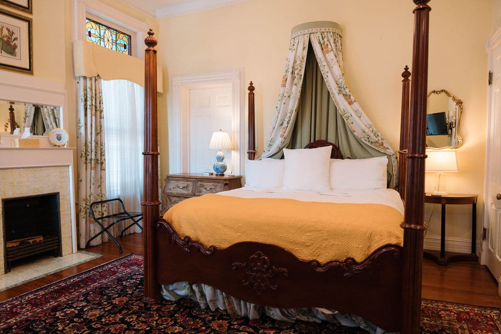 Bedroom suite featuring a dark wood four-poster bed with a canopy, a yellow throw blanket, a white fireplace, and a stained glass transom window.