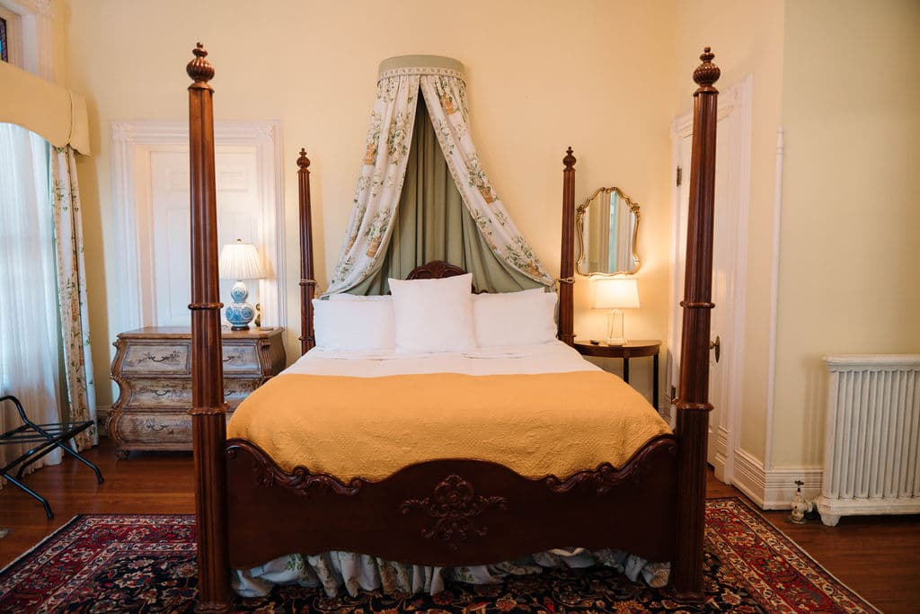 Straight-on view of an elegant bedroom suite featuring a dark wood four-poster bed with a floral canopy, gold throw blanket, antique chest, and Persian rug.