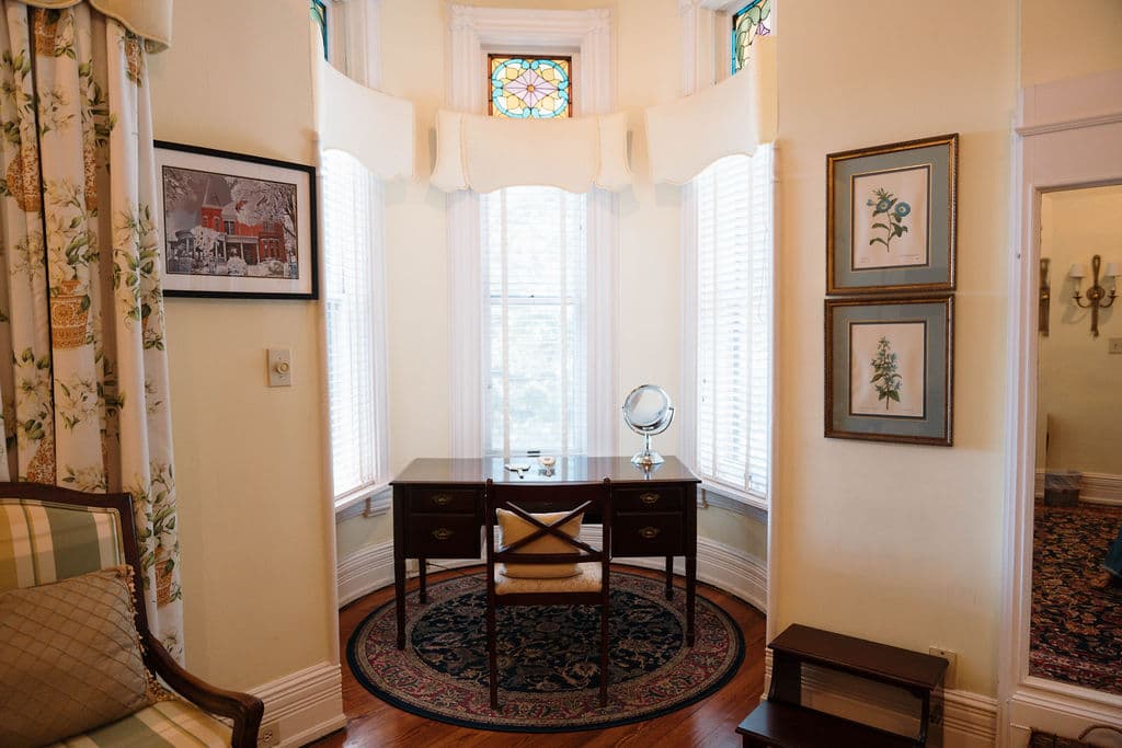 Sunny writing nook featuring a dark wooden desk placed in a bay window with stained glass transoms and framed artwork.