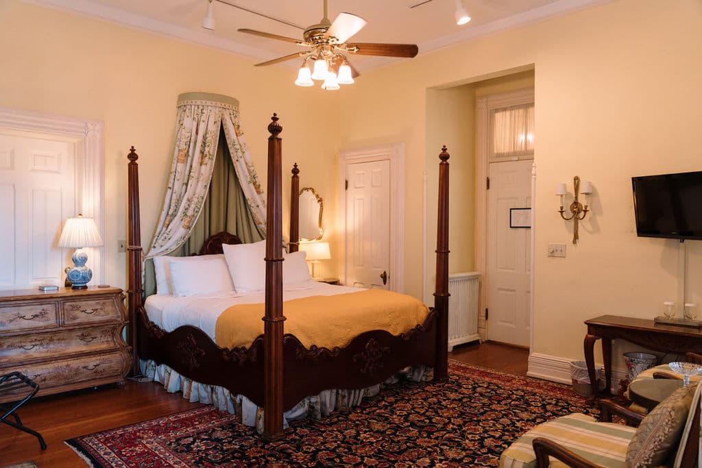 Spacious bedroom suite featuring a dark wood four-poster canopy bed, Persian rug, wall-mounted TV, and high ceilings.
