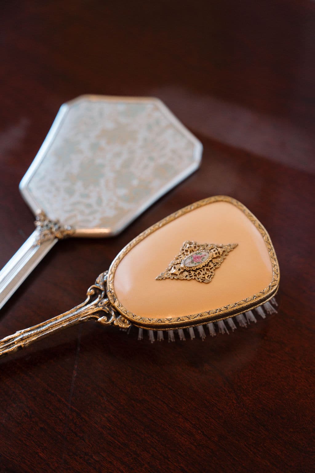 Close-up detail of an antique vanity set featuring a silver hand mirror and a matching brush with peach enamel backing sitting on a dark wood surface.