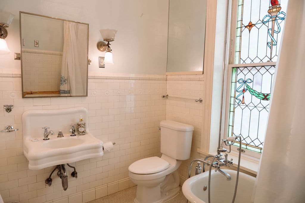 Bright, vintage-style bathroom suite featuring white subway tiles, a wall-mounted sink, and a large stained glass window next to the bathtub.