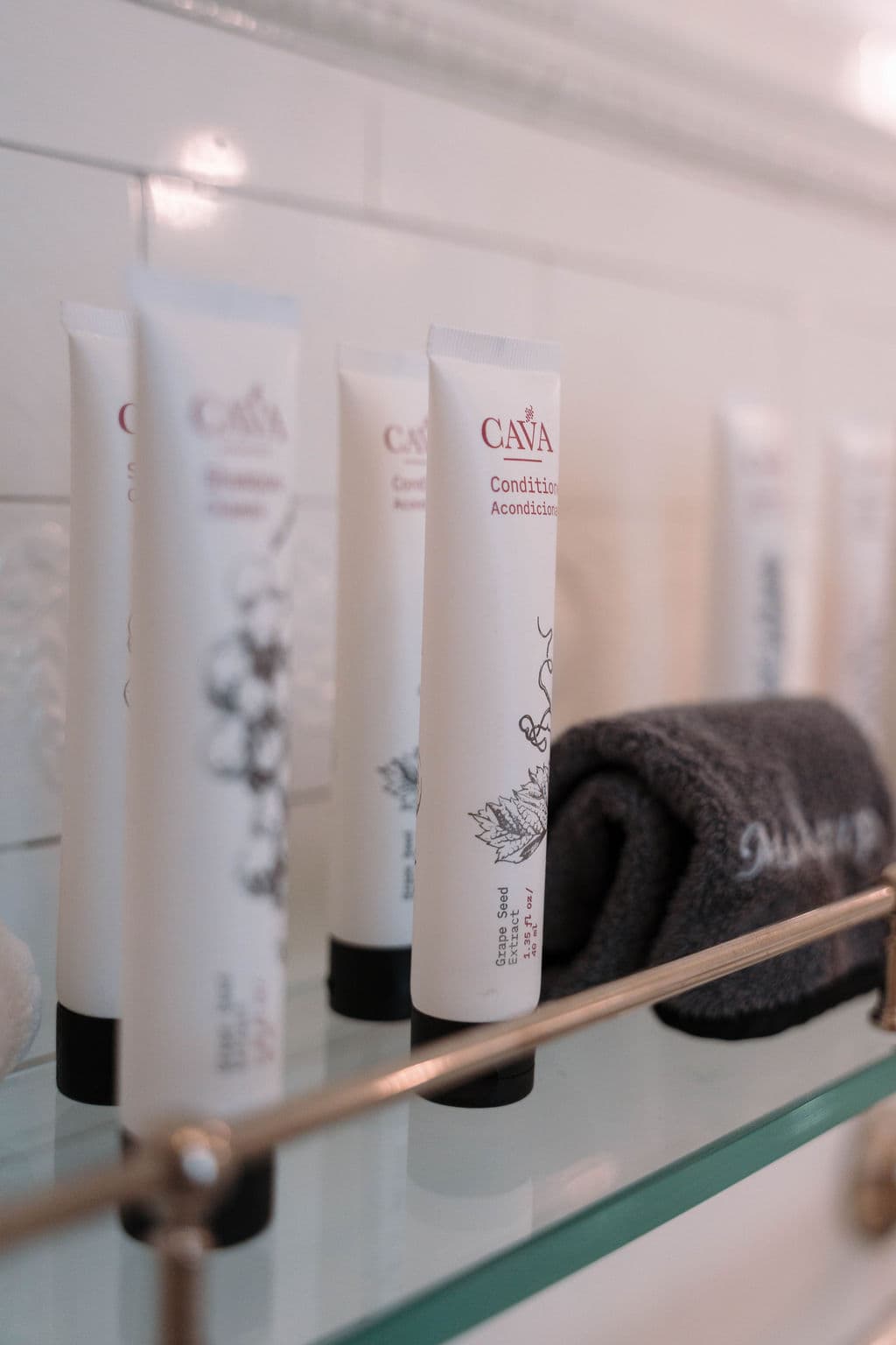 Close-up detail of premium bath amenity tubes and a rolled dark gray washcloth arranged on a glass shelf with brass railing against white subway tiles.
