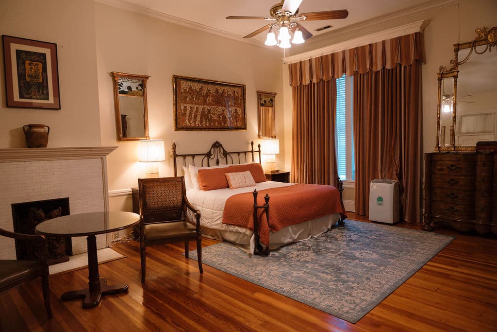 Spacious bedroom suite featuring hardwood floors, an iron bed with orange accents, a sitting area with a fireplace, and a large wooden dresser.