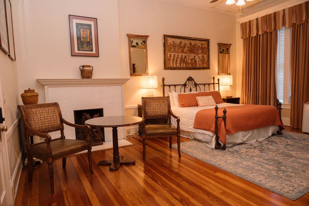 Bedroom suite featuring gleaming hardwood floors, a sitting area with two chairs by a white fireplace, and an iron bed with orange accents.