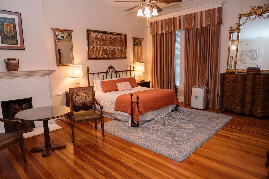 Spacious bedroom suite featuring hardwood floors, an iron bed with orange accents, a sitting area with a fireplace, and a large wooden dresser.