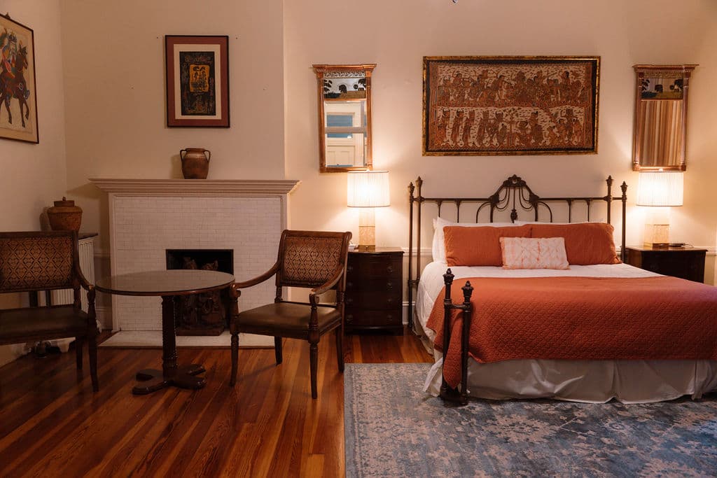 Bedroom suite featuring polished hardwood floors, a sitting area with two chairs by a white fireplace, and a black iron bed with orange bedding.