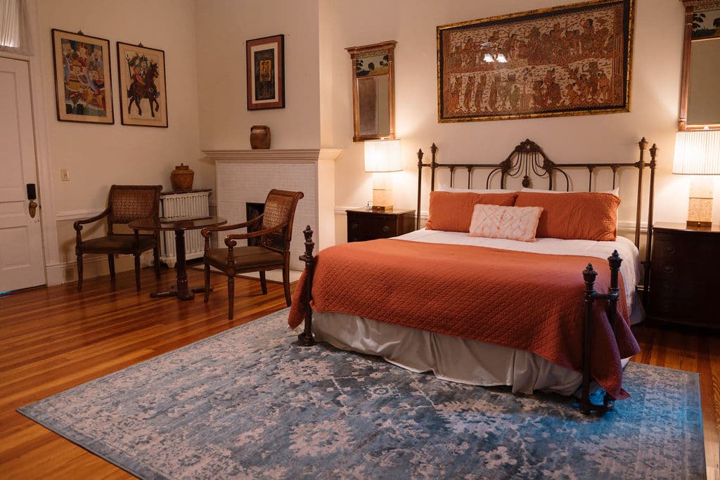 Bedroom suite featuring polished hardwood floors, a sitting area with two chairs by a white fireplace, and a black iron bed with orange bedding.
