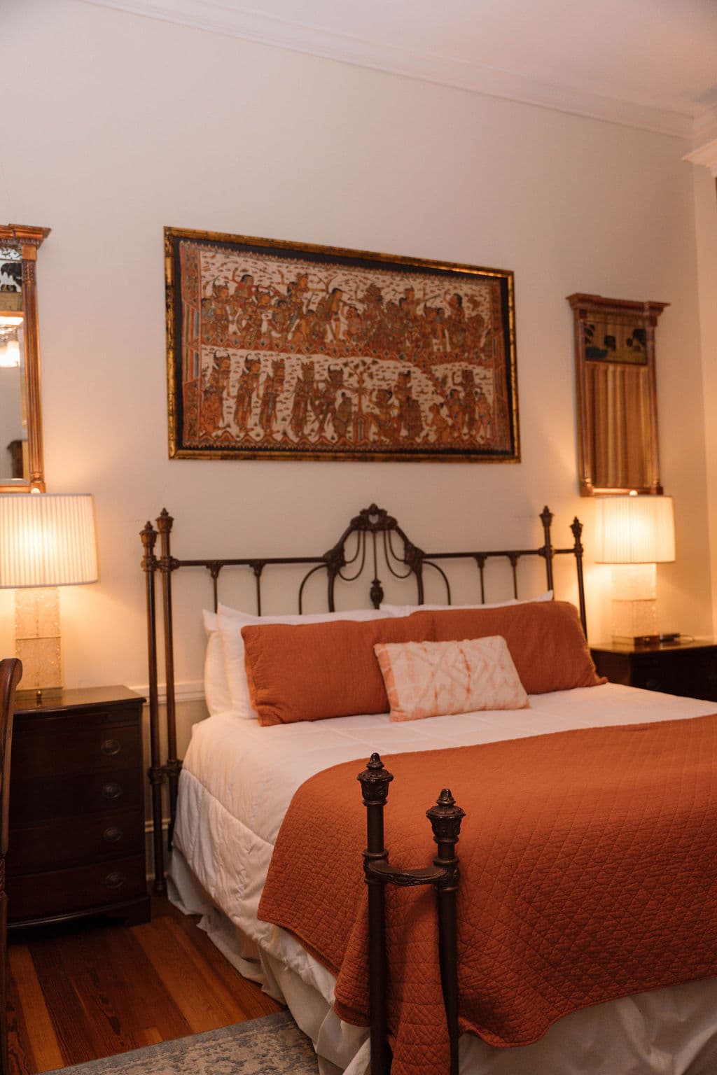 Close-up view of a black iron bed with rust-colored bedding and pillows, flanked by warm bedside lamps and artwork hanging above the headboard.