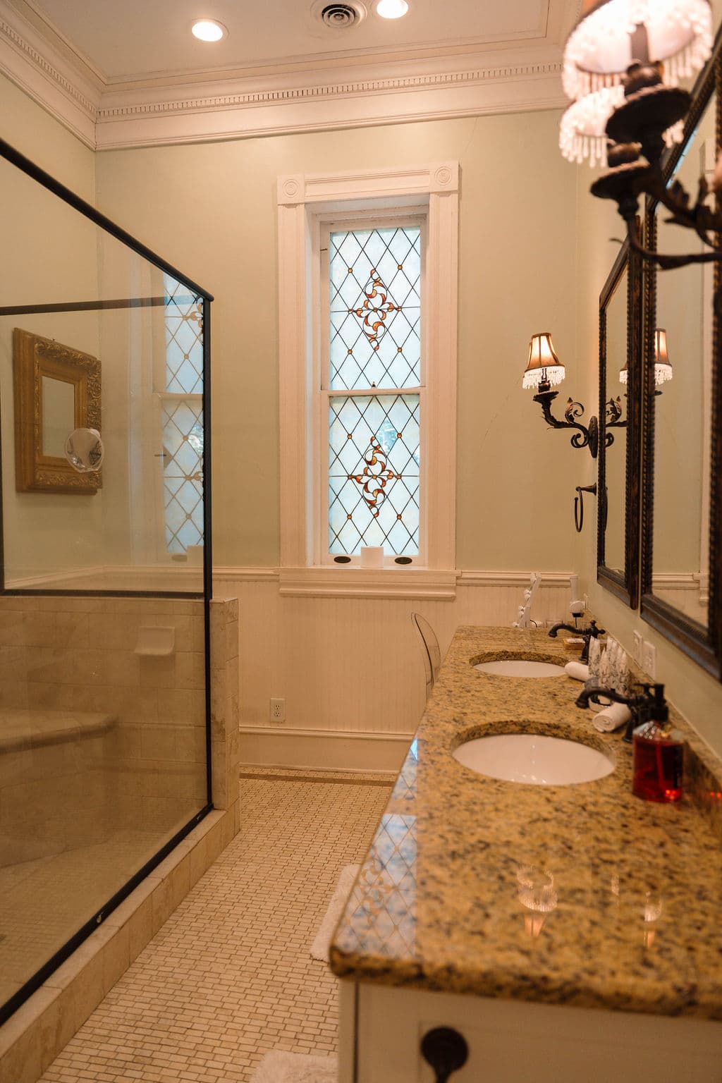 Spacious bathroom suite featuring a granite double vanity, a glass-enclosed walk-in shower, and a tall diamond-patterned stained glass window.