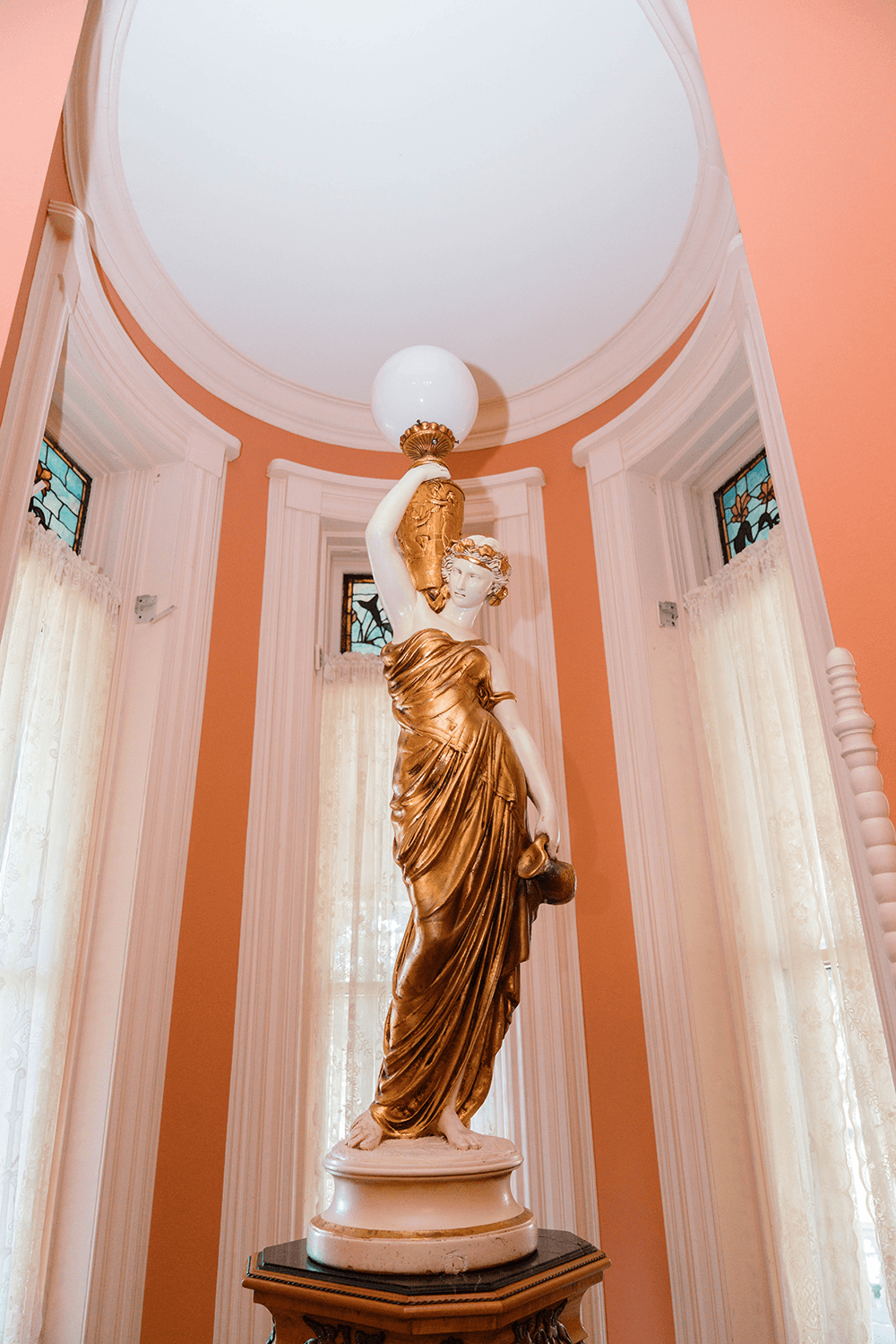 A gold and white statue of a woman holding a lamp stands in a room with orange walls and stained glass windows.