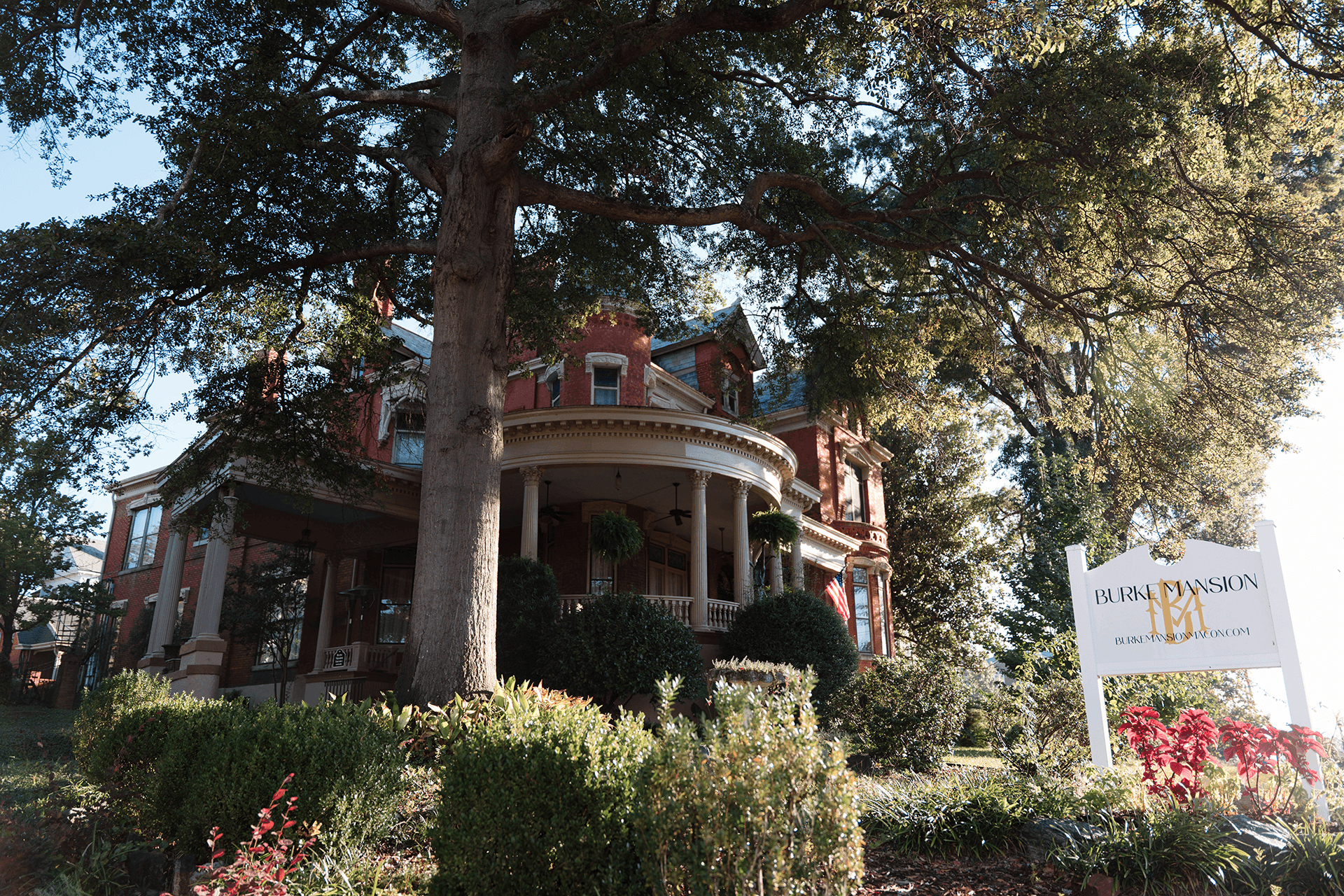 A grand, red-brick Victorian mansion with large windows and a circular porch. Lush greenery and a tree frame it, with a sign reading "Park Mansion" in front.