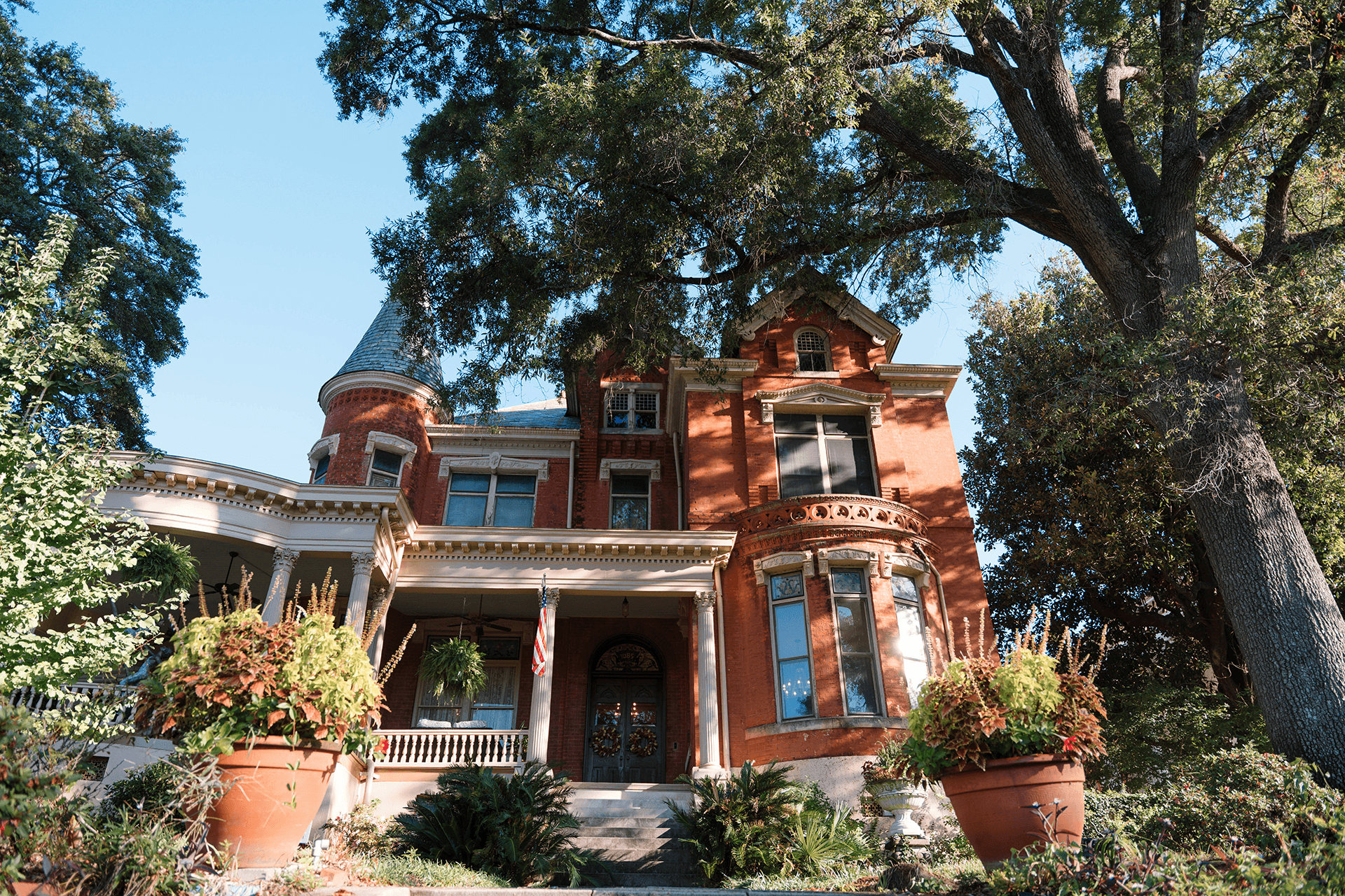 A grand red brick house with intricate architecture, nestled among large trees and colorful potted plants.