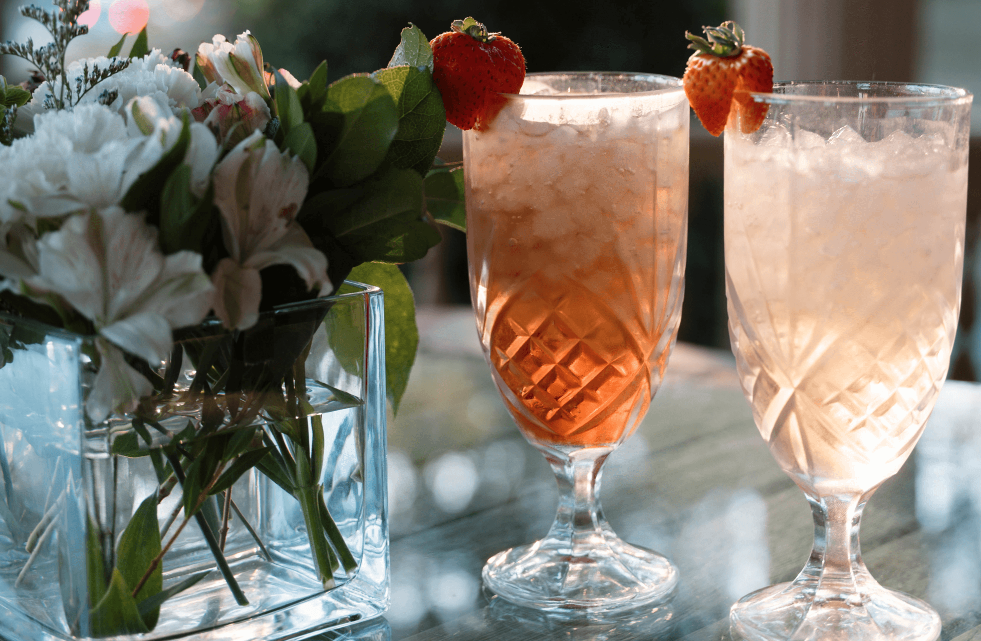 Two crystal glasses filled with iced drinks and garnished with strawberries, placed next to a flower arrangement.