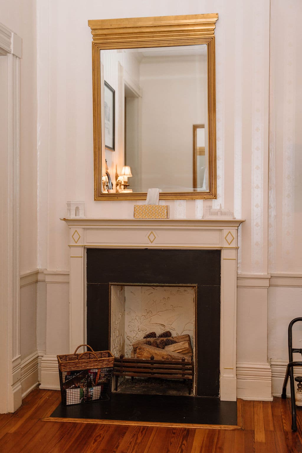 White fireplace mantel with a black surround and hearth, featuring a large rectangular gold-framed mirror hanging above it. A wire magazine basket sits on the hardwood floor to the left of the fireplace.