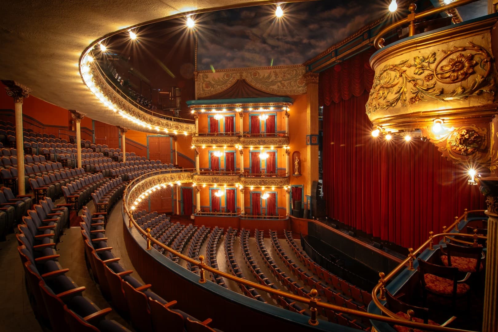 Opulent theater interior with curved seating, ornate balconies, and red curtains. Soft, warm lighting creates a grand and inviting atmosphere.
