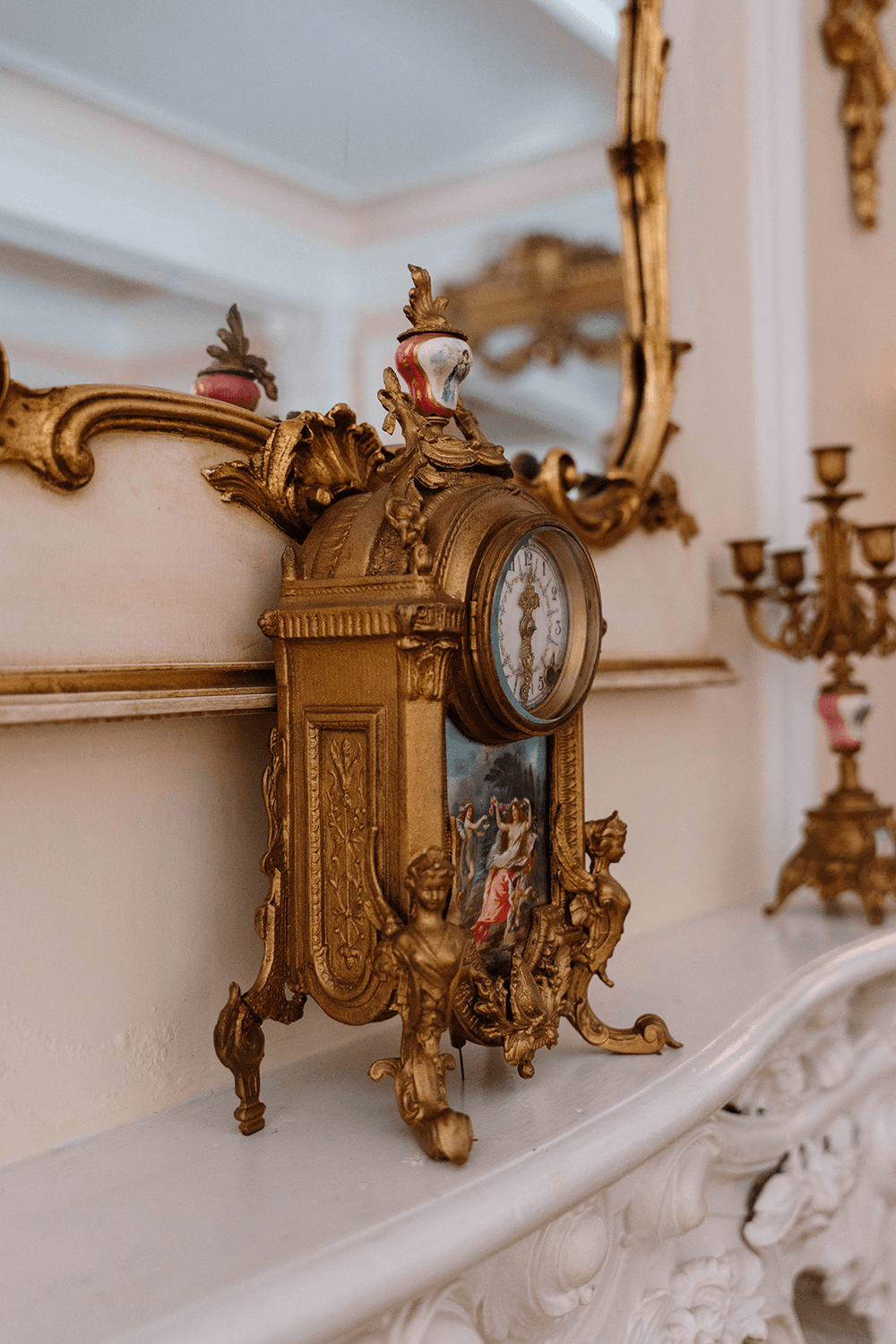 An ornate gold mantel clock sits on a white ledge, reflecting a decorative mirror in the background.