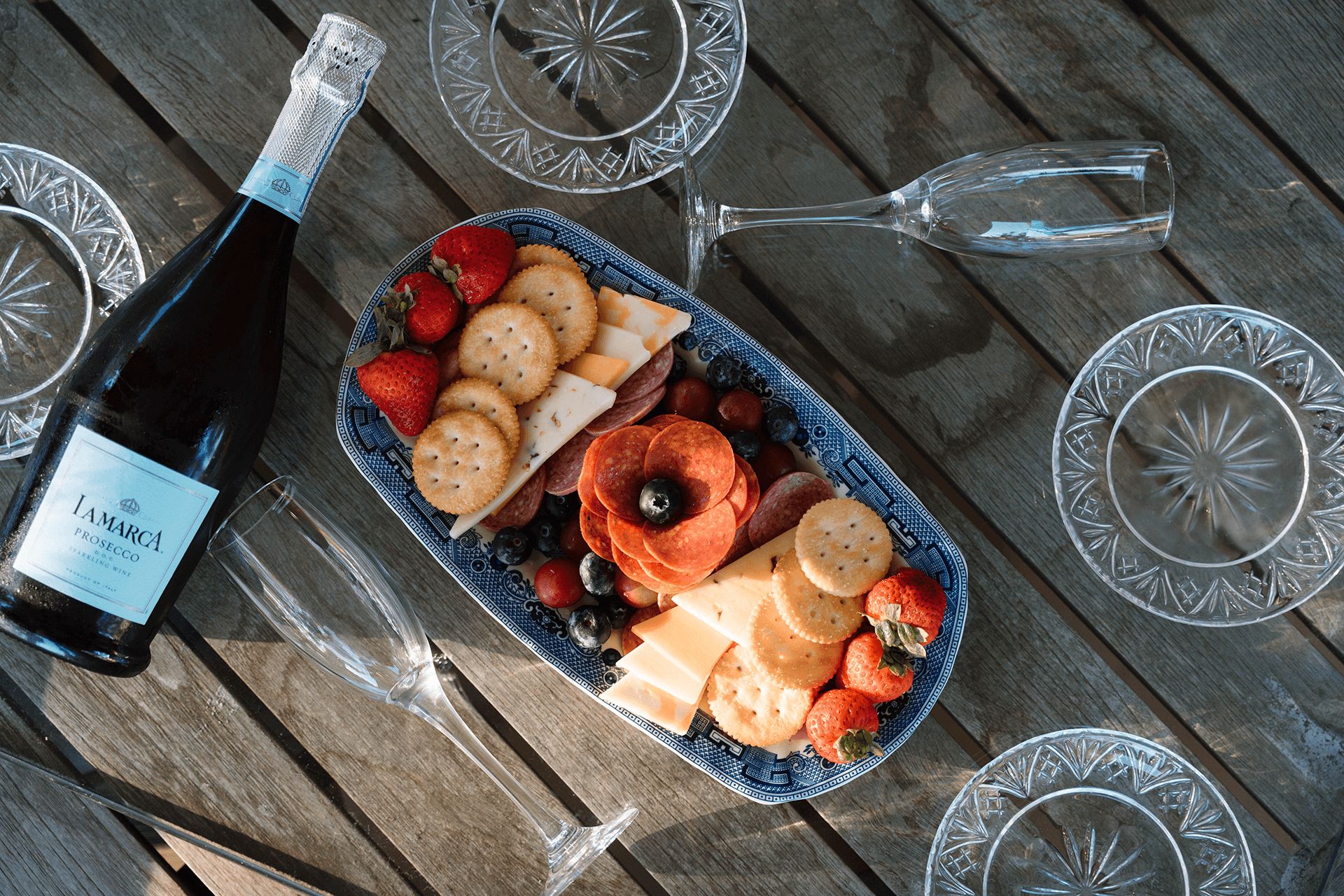 A wooden table with a blue platter of cheese, crackers, strawberries, blueberries, and salami. A bottle of Prosecco and empty glasses surround it.