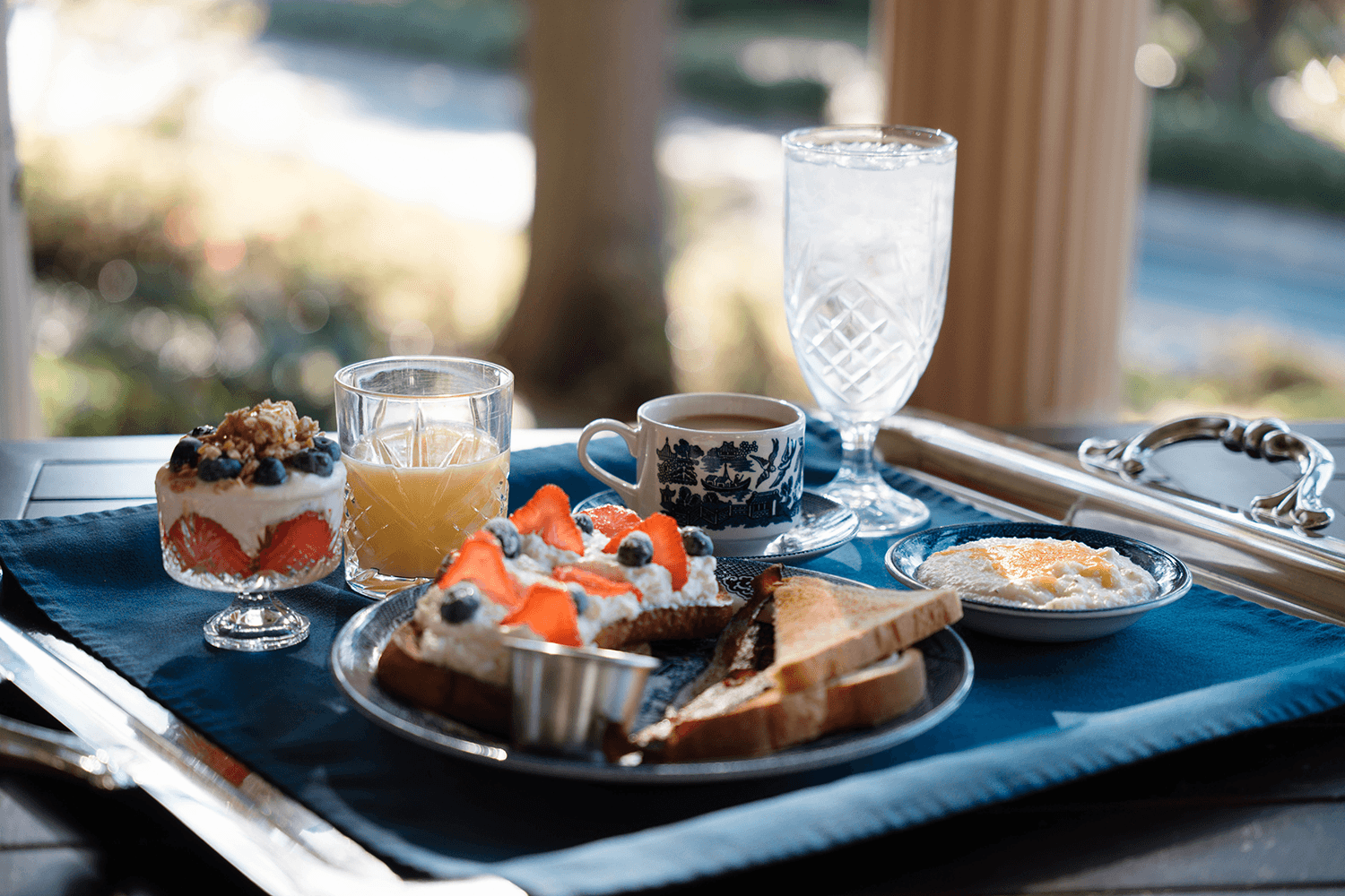A beautifully arranged breakfast spread featuring toast, fresh fruit, yogurt, juice, coffee, and water on a blue tablecloth.
