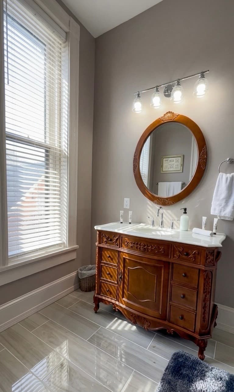 A bright bathroom featuring a vintage wooden vanity, round mirror, and large window with blinds.