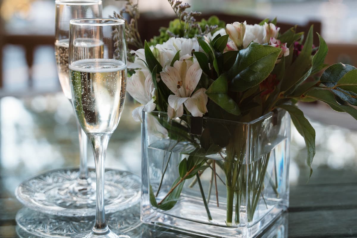 Two flutes of sparkling wine beside a floral arrangement in a glass vase on a table.