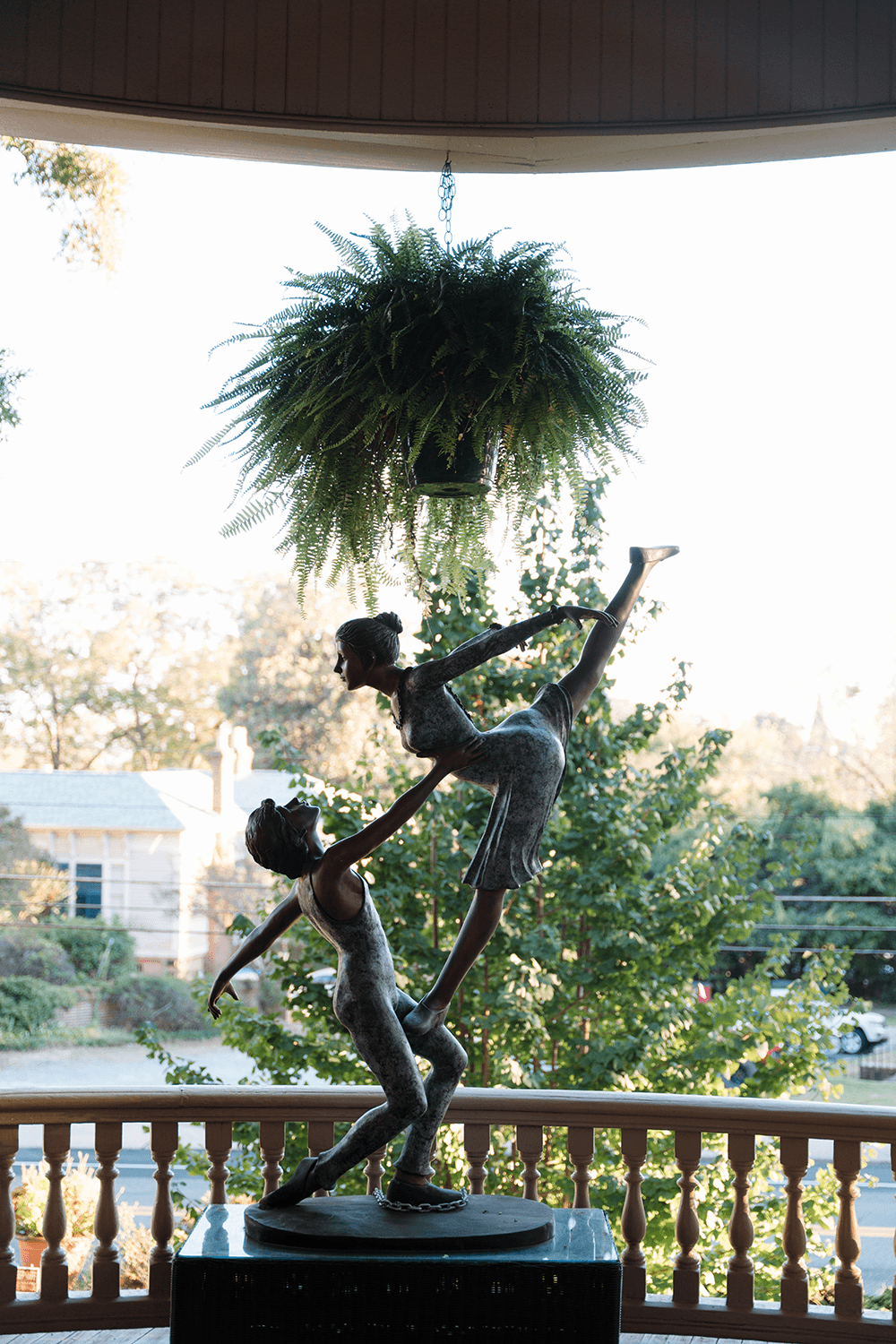 A bronze sculpture of two dancers is framed by a hanging plant and greenery outside an ornate porch.