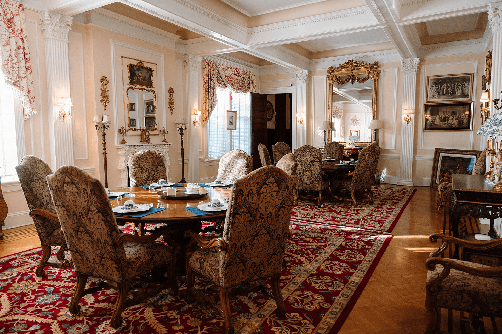 Elegant dining room with ornate chairs around a wooden table set with teacups. Decor includes large mirrors, intricate carpeting, and classical artwork, creating a luxurious and inviting atmosphere.