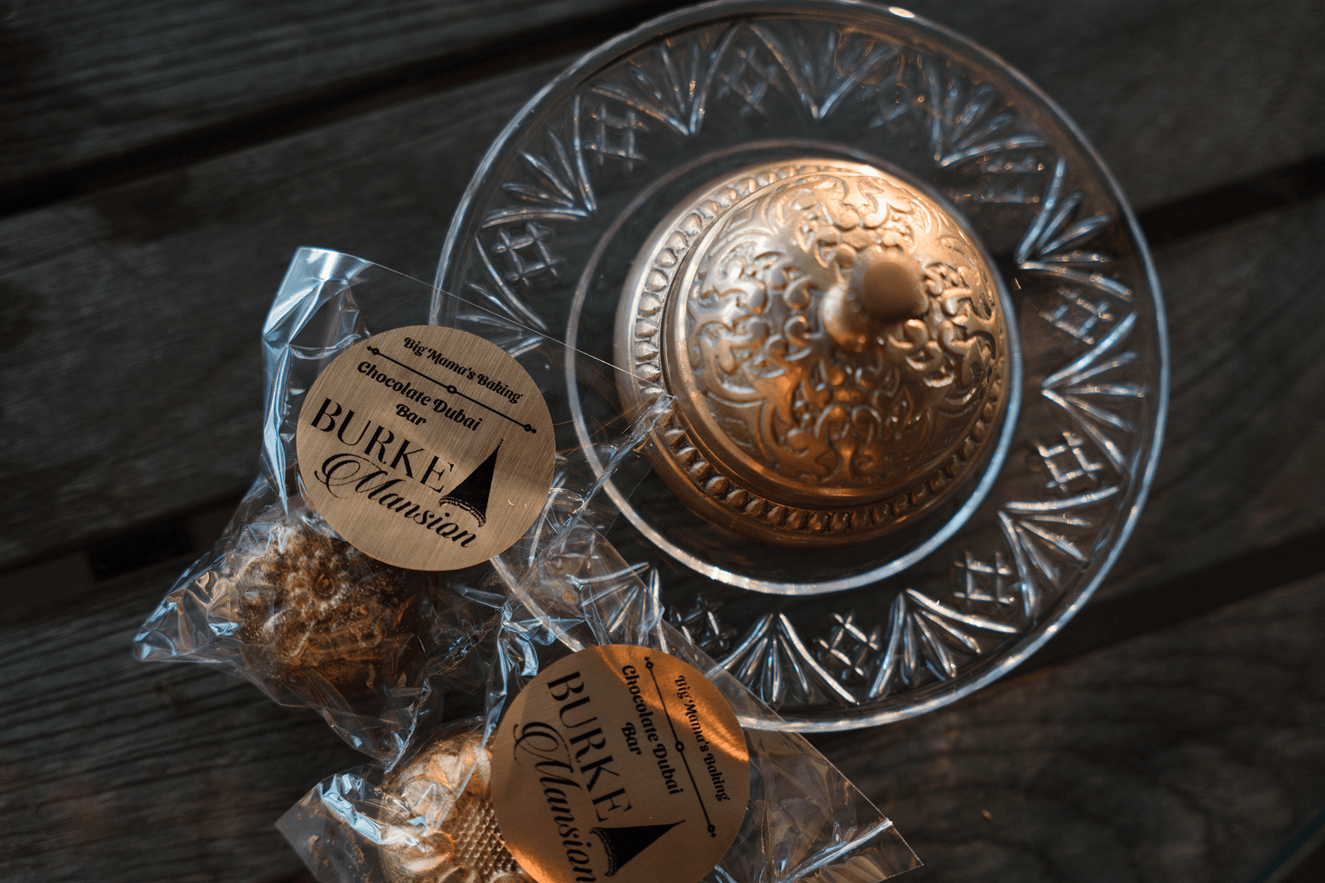 Elegant glass dish with ornate, golden lid on wood surface. Nearby, wrapped treats labeled "BURKE Mansion" exude luxury and warmth.