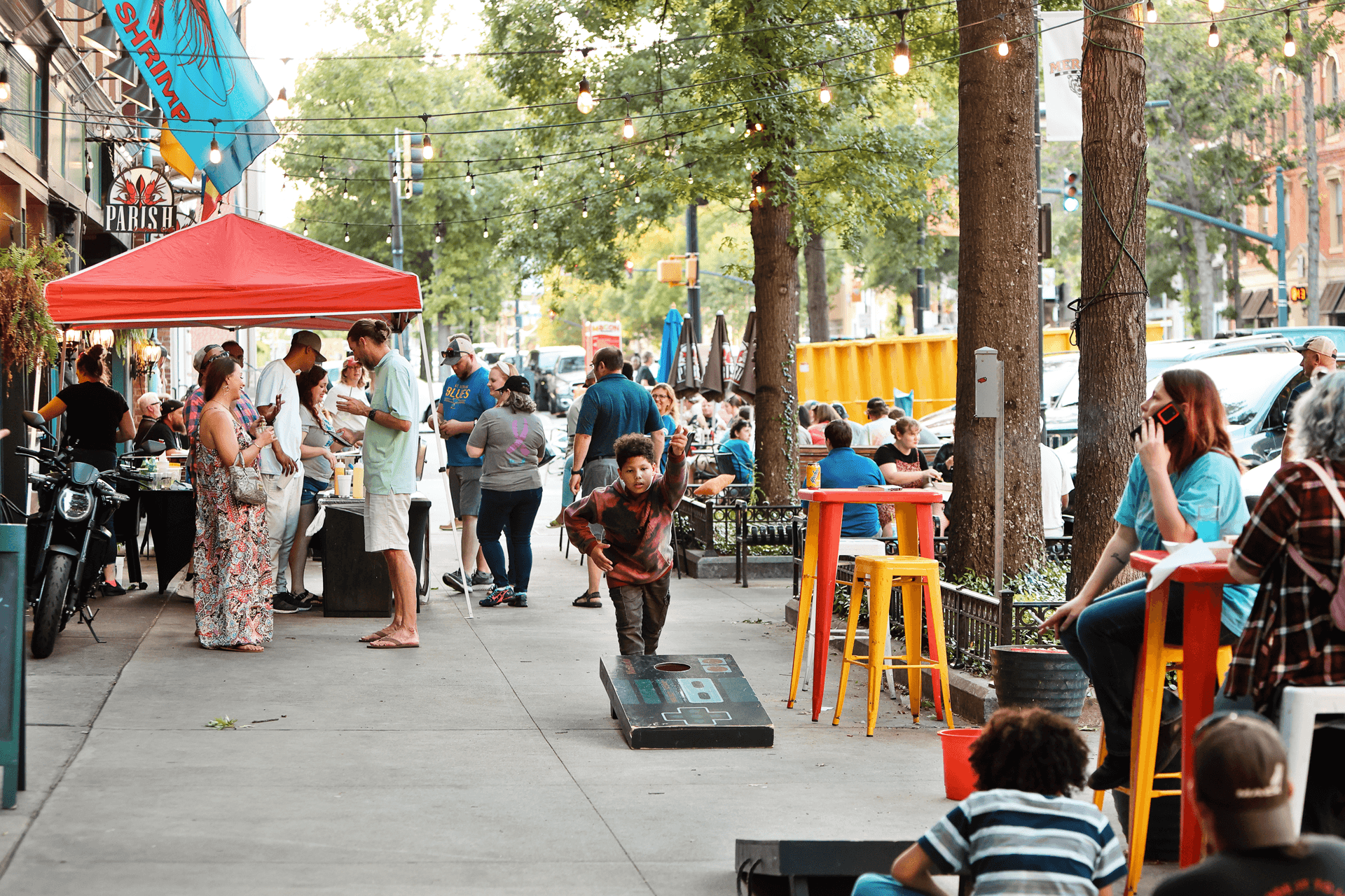 A bustling outdoor scene with people socializing, dining, and a child playing a bean bag toss game on a sunny day.