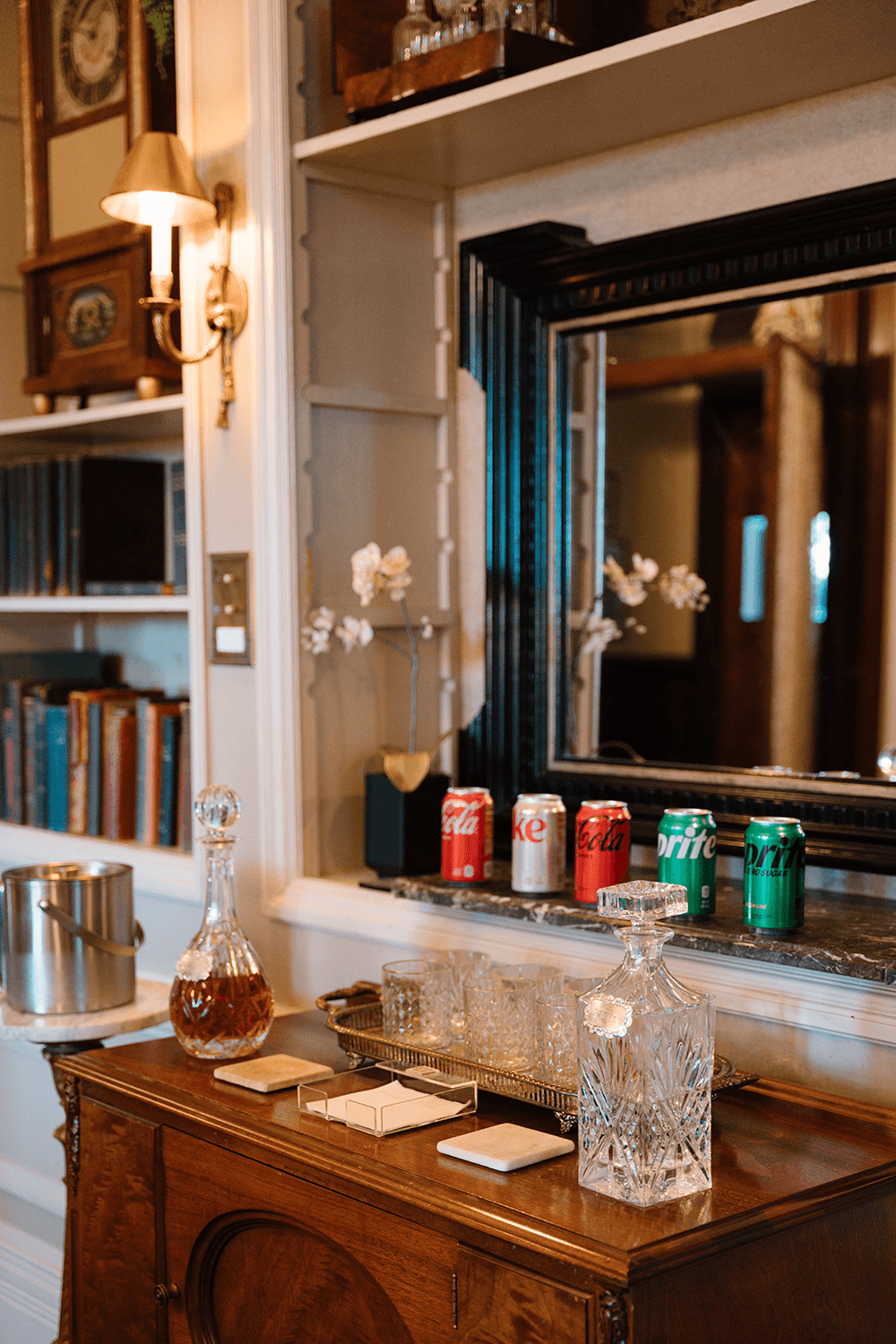 Elegant room with a wooden sideboard holding glass decanters, coasters, ice bucket, and assorted soda cans. A mirror reflects the cozy, sophisticated atmosphere.