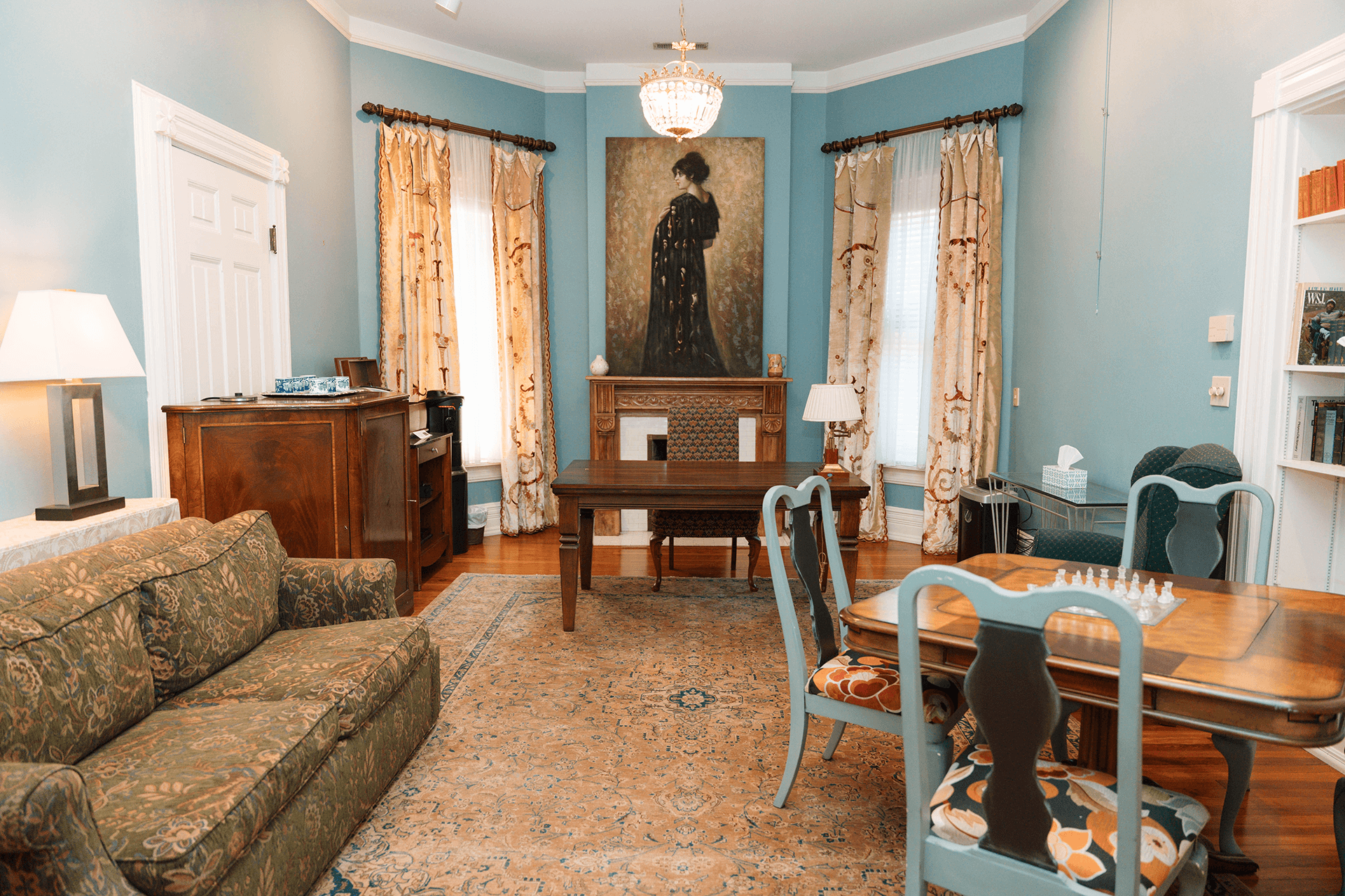 A cozy room featuring a green sofa, a wooden table, a painting of a woman, and decorative curtains.