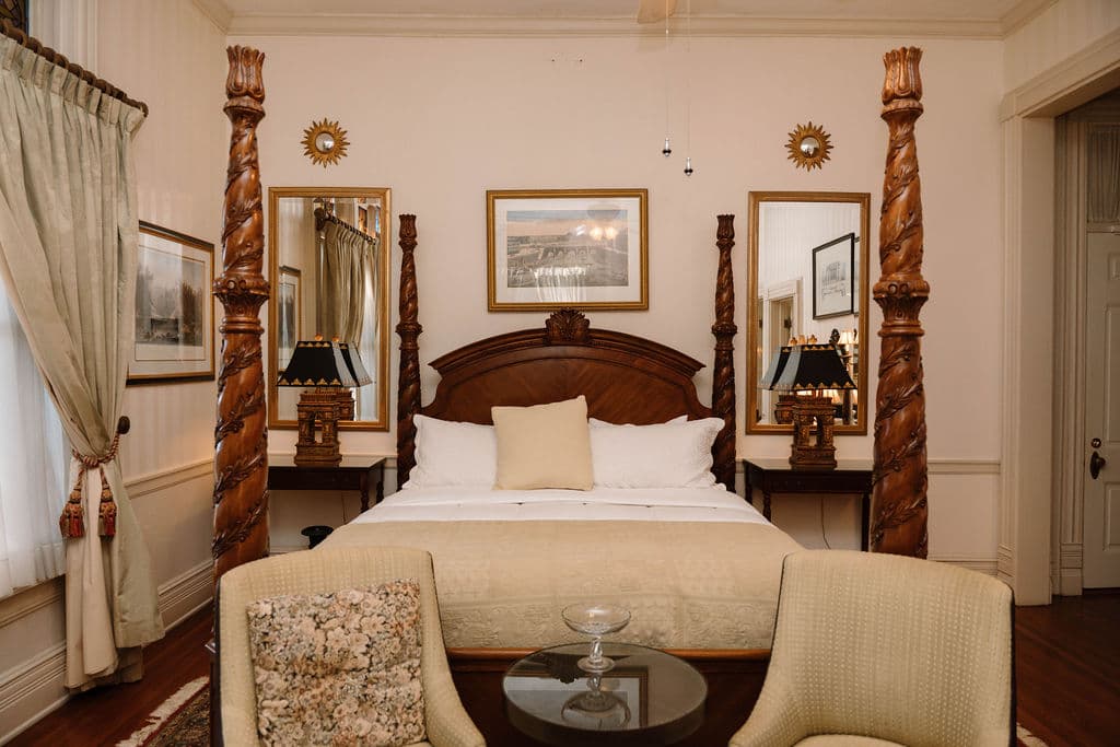 A classic four-poster bed with spiral posts, flanked by tall mirrors and nightstands, with two beige armchairs at the foot of the bed.